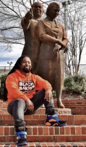 Photo of Trey Benham in front of a statue in an orange hoodie that reads "Black Bartow" across the chest. 